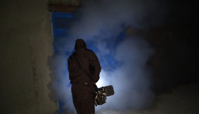 کارگر مجهز به لباس ایمنی در حال انجام فرآیند مه‌پاشی برای کنترل آفات و ضدعفونی محیط صنعتی در ساعات شب.