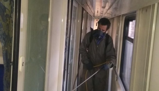 کارگر با لباس ایمنی و ماسک محافظ در حال انجام عملیات سم‌پاشی و ضدعفونی در راهروی یک واگن قطار، به منظور کنترل آفات و افزایش بهداشت محیطی.